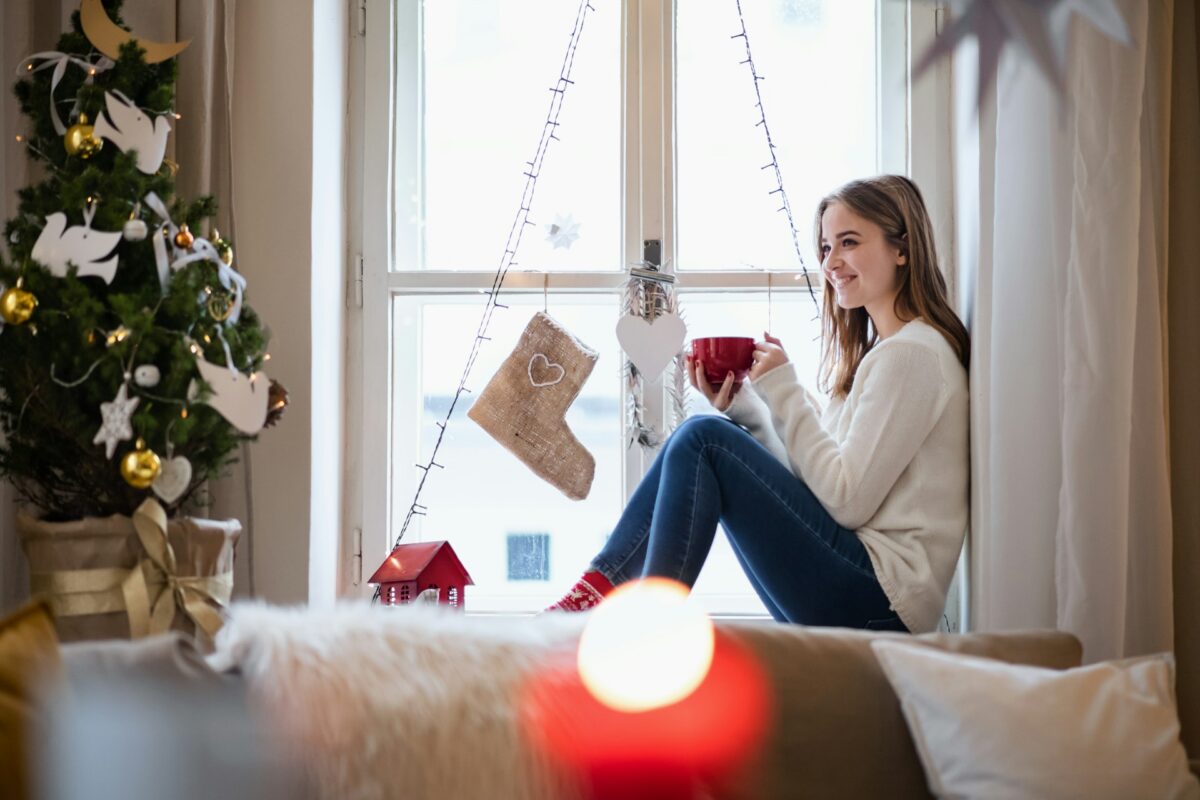 Woman relaxing by a window with a warm drink during cosmetic surgery recovery in winter.