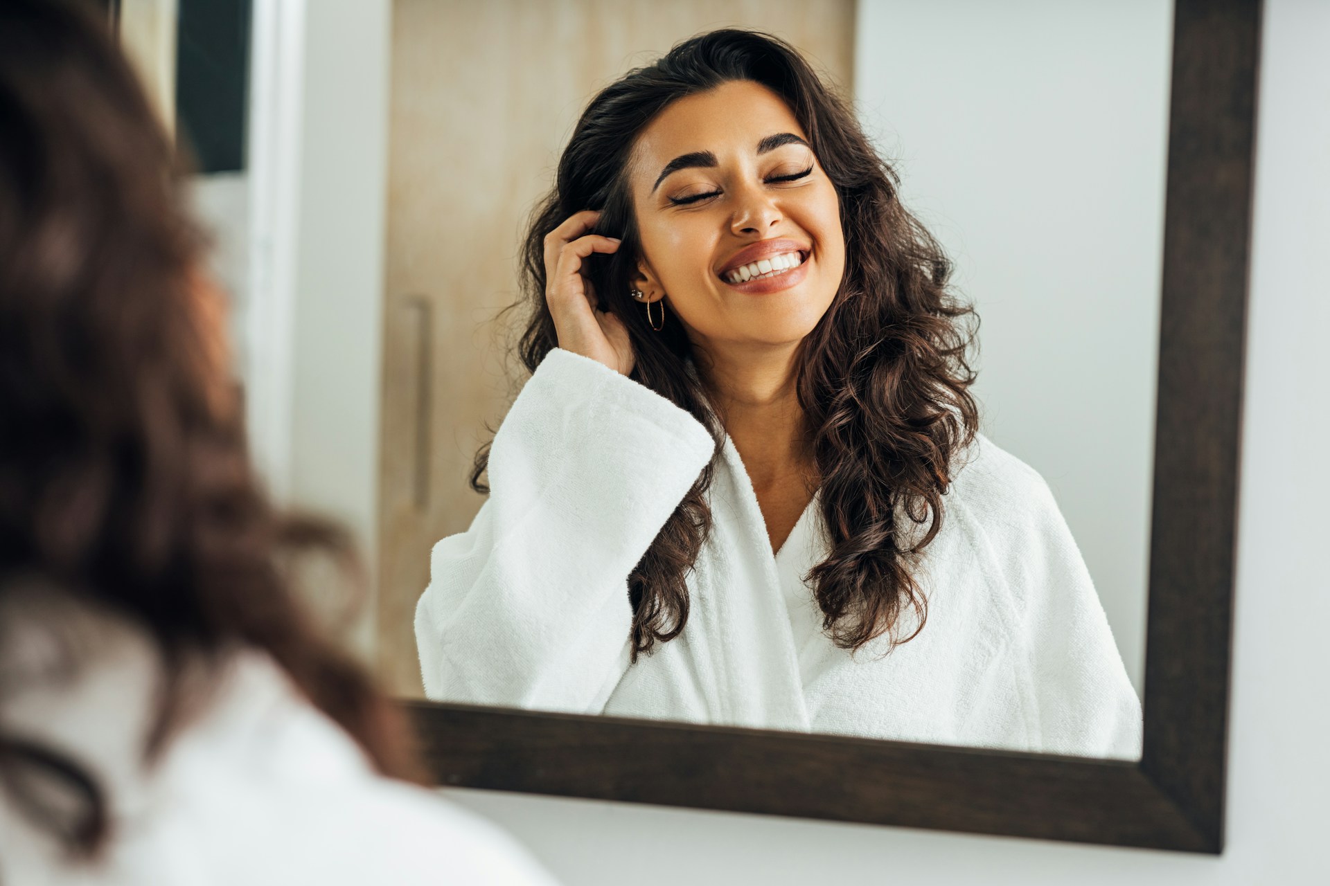Woman smiling at her reflection while preparing for cosmetic surgery and feeling confident about her aesthetic goals.